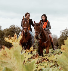 Two riders on horses, one horse has a mouthful of grass