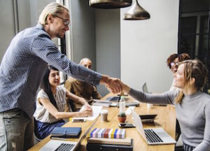 A business meeting, four people sitting down, one man standing, shaking the hand of a woman, everyone is smiling